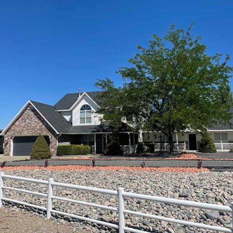 Suburban house with stone facade, front yard landscaping, and a white fence under a clear blue sky