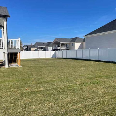 Backyard with green lawn, white privacy fence, and neighboring houses under a clear blue sky