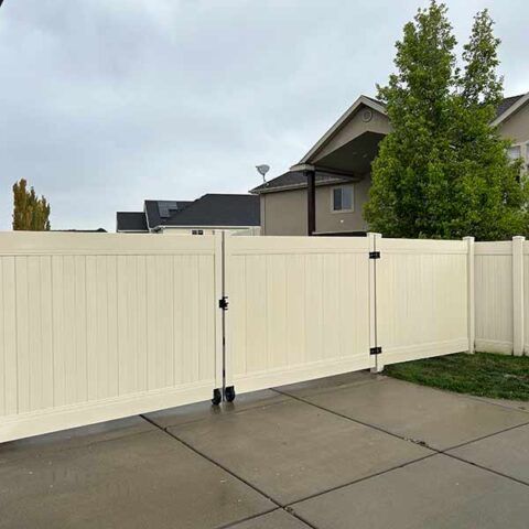 White vinyl fence with a double gate along a concrete driveway in a suburban yard.