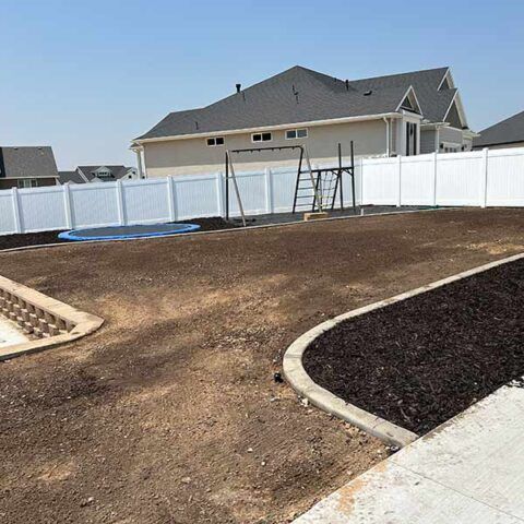 Freshly landscaped backyard with dirt, mulch, and a white vinyl fence under a clear sky