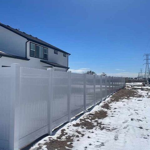 Snow-covered yard beside a house with a long white privacy fence under a clear blue sky