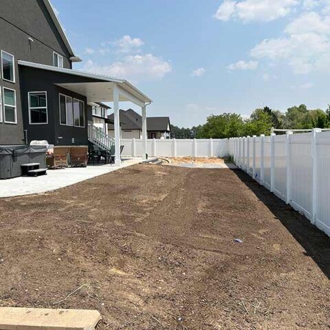 Fresh dirt backyard beside a gray house and white fence, with a covered patio and new landscaping area