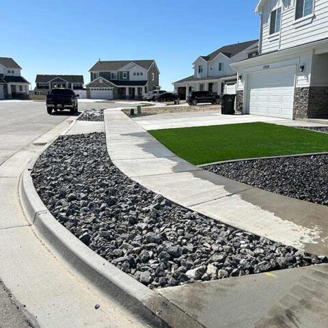 Suburban street corner with rock landscaping, concrete curbs, and a green lawn under a clear blue sky