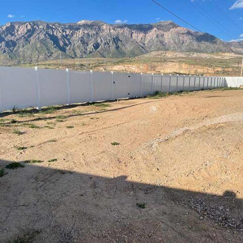 Dirt lot beside a white fence, with mountains under a clear blue sky in the background.