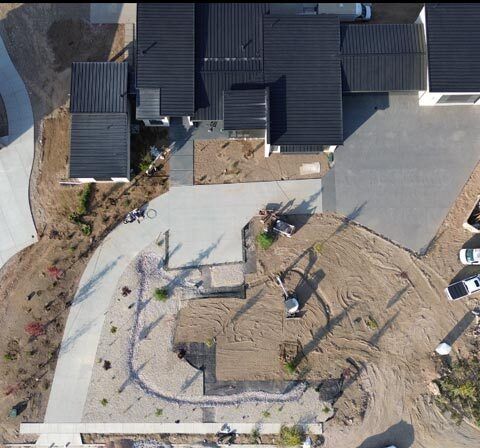 Aerial view of a house under construction with a concrete driveway, dirt lot, and parked vehicles nearby