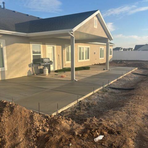 New concrete patio beside a tan house with a covered porch, viewed from a dirt yard.