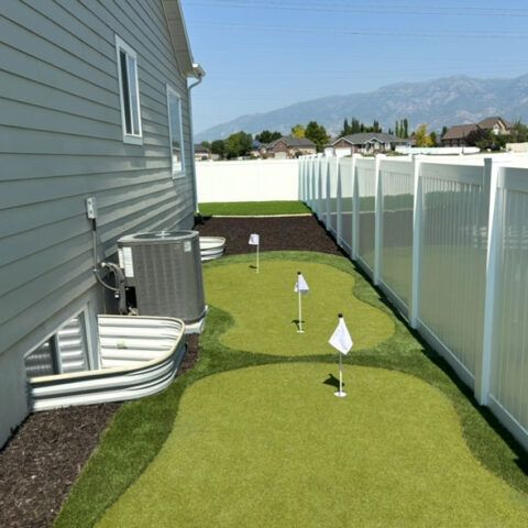 Narrow backyard with artificial turf putting green beside a house and white fence, with mountains in the distance