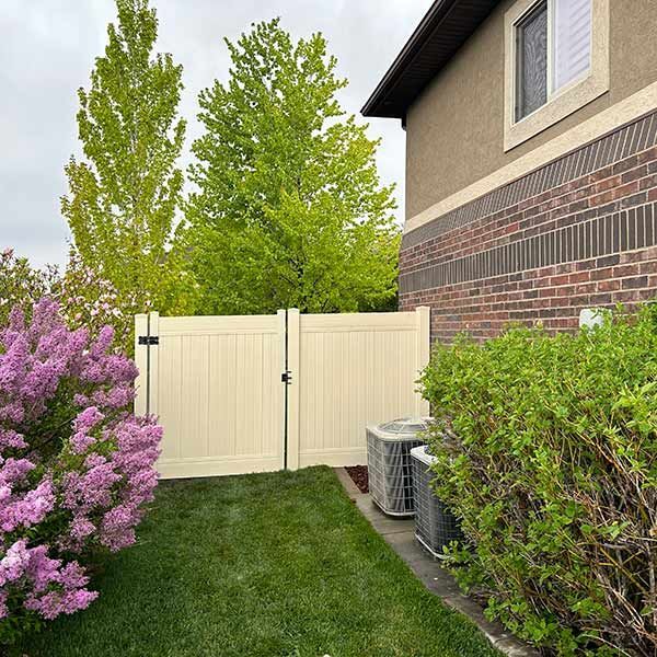 Backyard corner with white fence, green lawn, pink flowering shrub, and house exterior with air conditioner unit