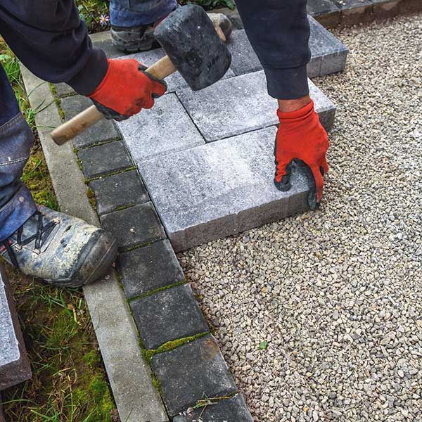 Worker laying gray paving slabs beside a gravel path, using a mallet and red gloves