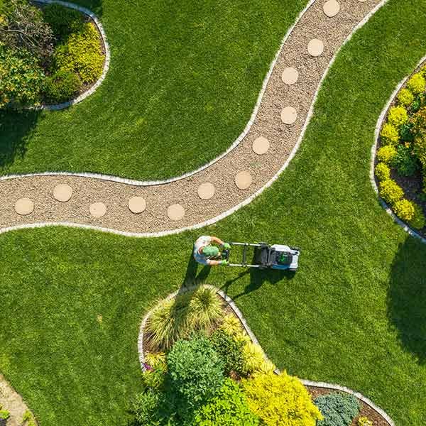 Overhead view of a winding garden path through green grass with a worker mowing near shrubs