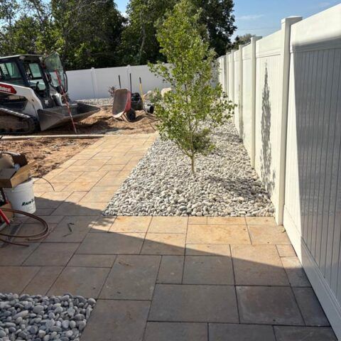 Backyard patio under construction with paver walkway, gravel beds, and a young tree along a white fence