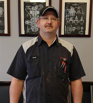 A technician in a black and grey work shirt with tools in his pocket, standing in front of two framed technical drawings.