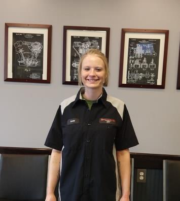 A person in a black and gray work uniform smiles in front of a wall with three framed engine patent prints.