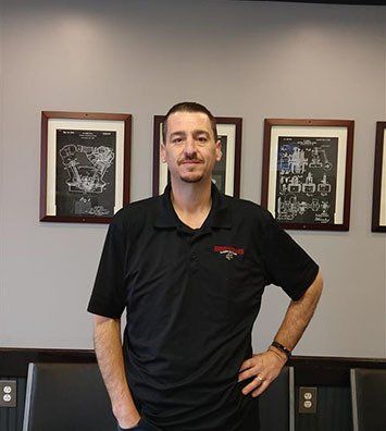 A person in a black polo shirt stands in an office in front of a wall with three framed technical drawings.