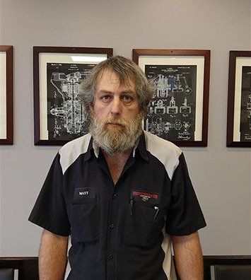 A bearded person wearing a two-tone black and grey work shirt stands before a wall with framed technical illustrations.
