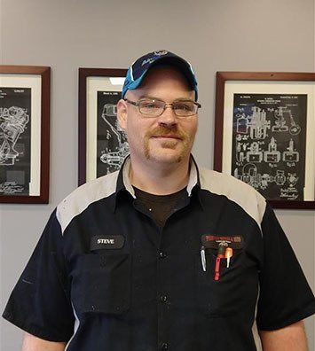 A person wearing a two-tone work shirt, glasses, and a cap stands centered in front of three framed technical diagrams.