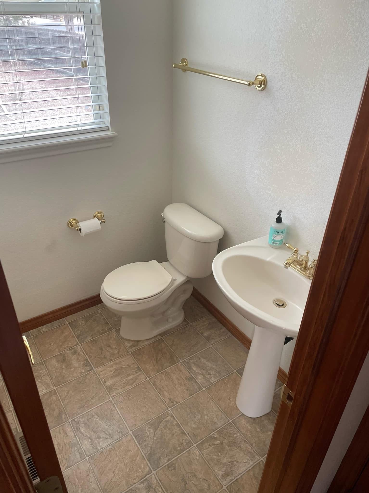 Small beige bathroom with toilet, pedestal sink, towel bars, and a window with blinds.