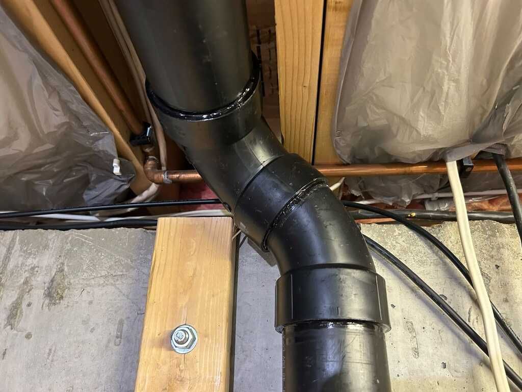 Black sewer pipe elbow in attic between joists, with insulation and wiring nearby.