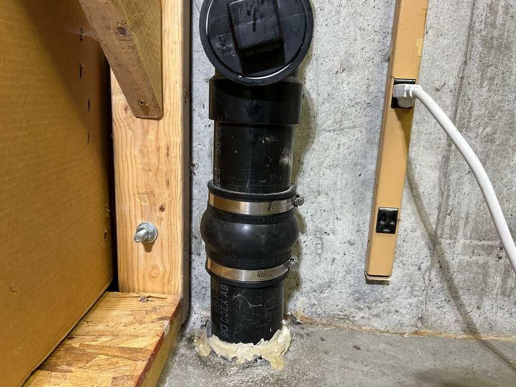 Black vertical plumbing pipe with fittings beside a concrete wall and wooden framing.