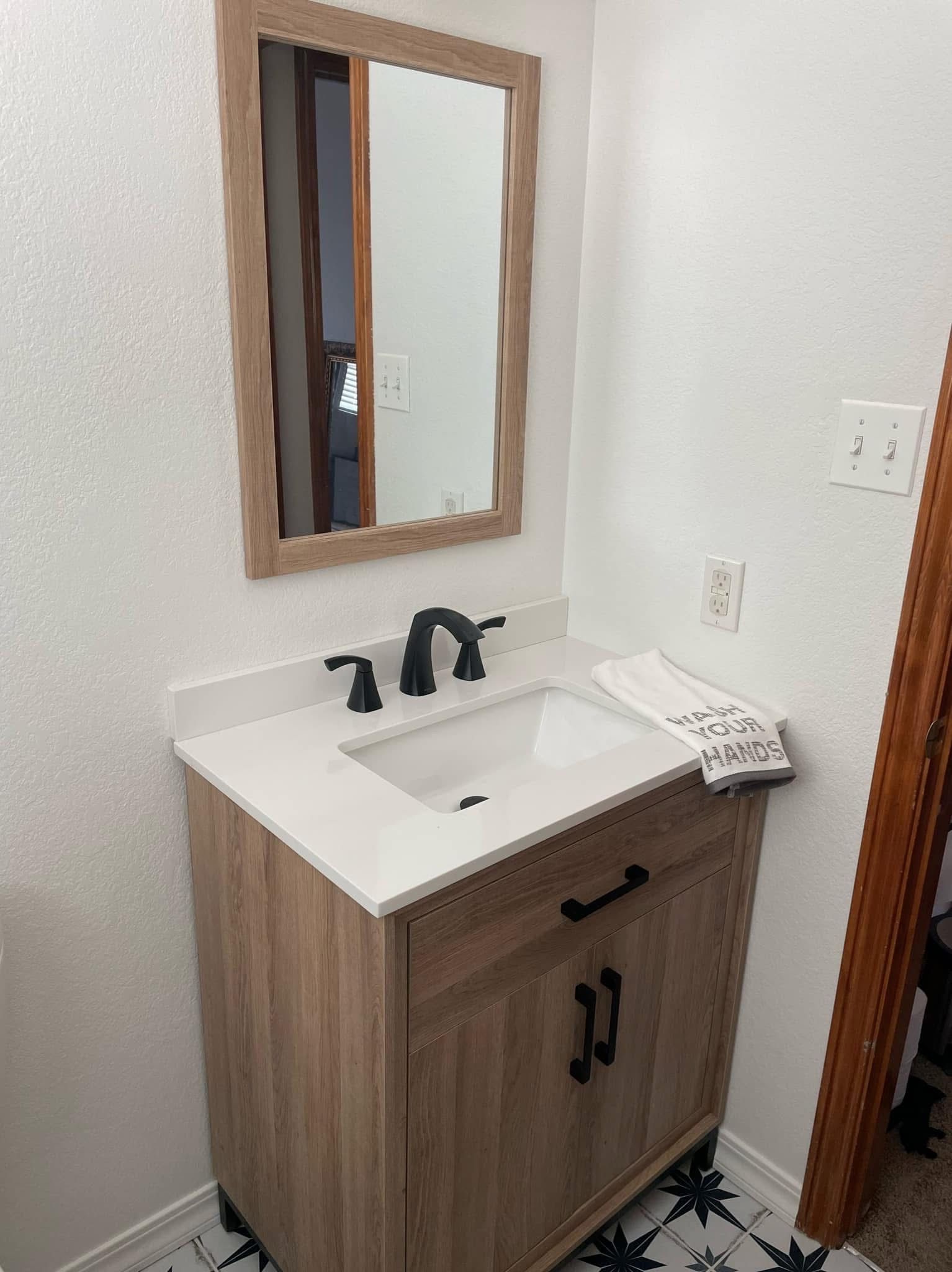 Small bathroom vanity with wood cabinet, white sink, black faucet, and framed mirror.