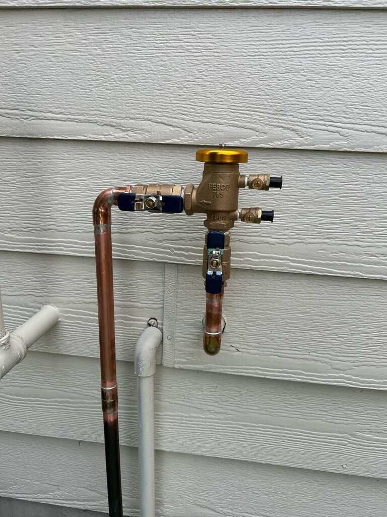 Outdoor copper plumbing pipe assembly mounted on gray house siding with a yellow valve handle.