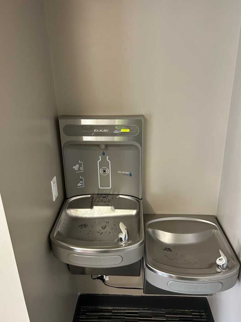 Wall-mounted stainless steel water fountain and pet bowl in a corner of a plain room.