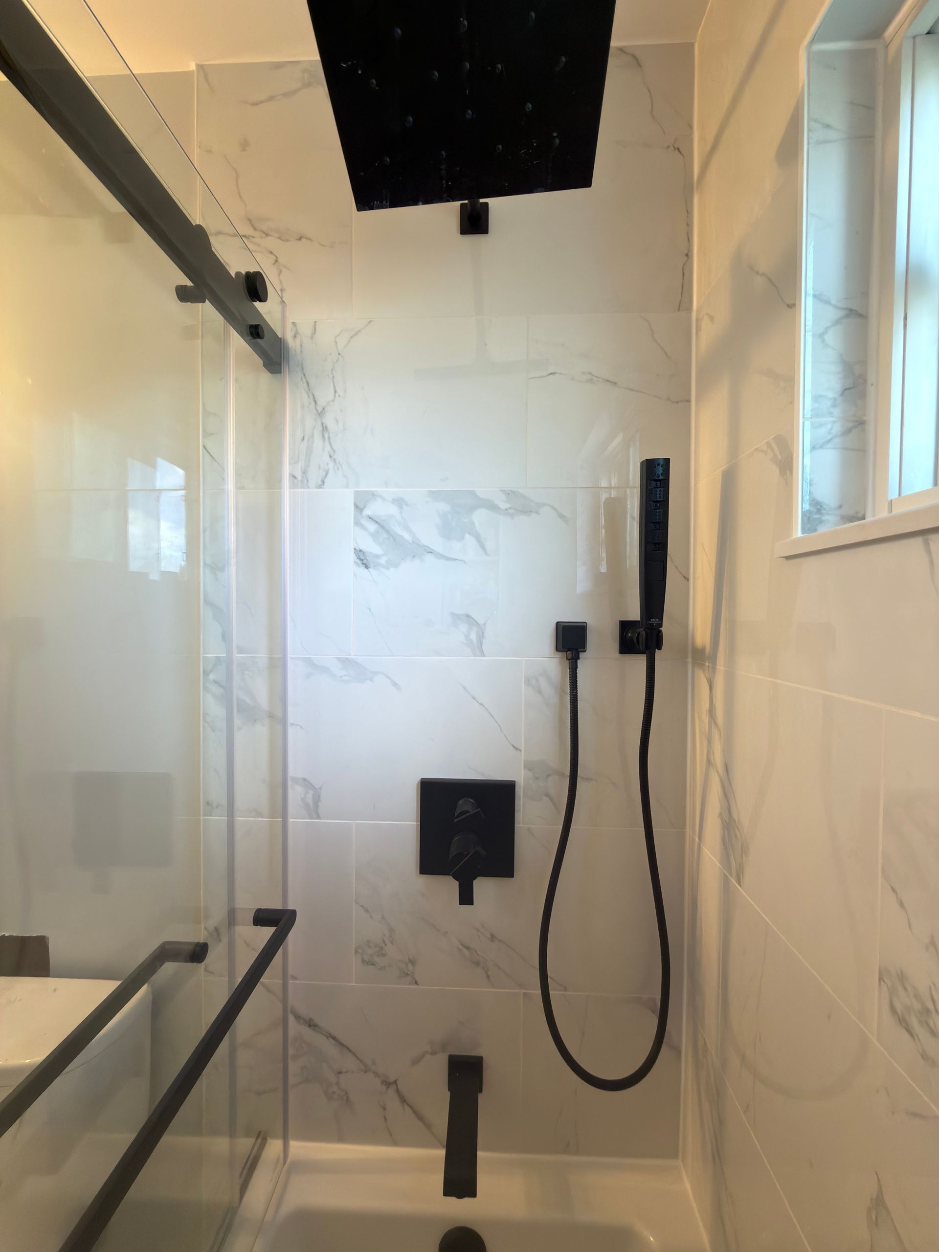 Modern shower with white tile, glass door, black rain shower head, and handheld sprayer.