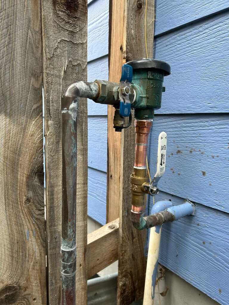 Weathered outdoor plumbing with copper pipe, valve, and faucet beside blue house siding.