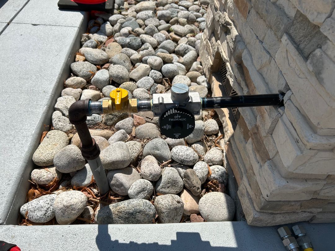 Outdoor utility pipes and valves set in a rock bed beside a stone wall and concrete walkway.