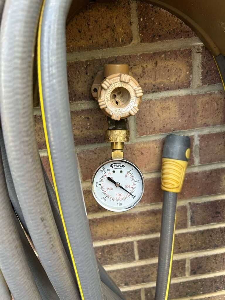 Wall-mounted hose bib with pressure gauge and black-and-yellow hose against a brick wall