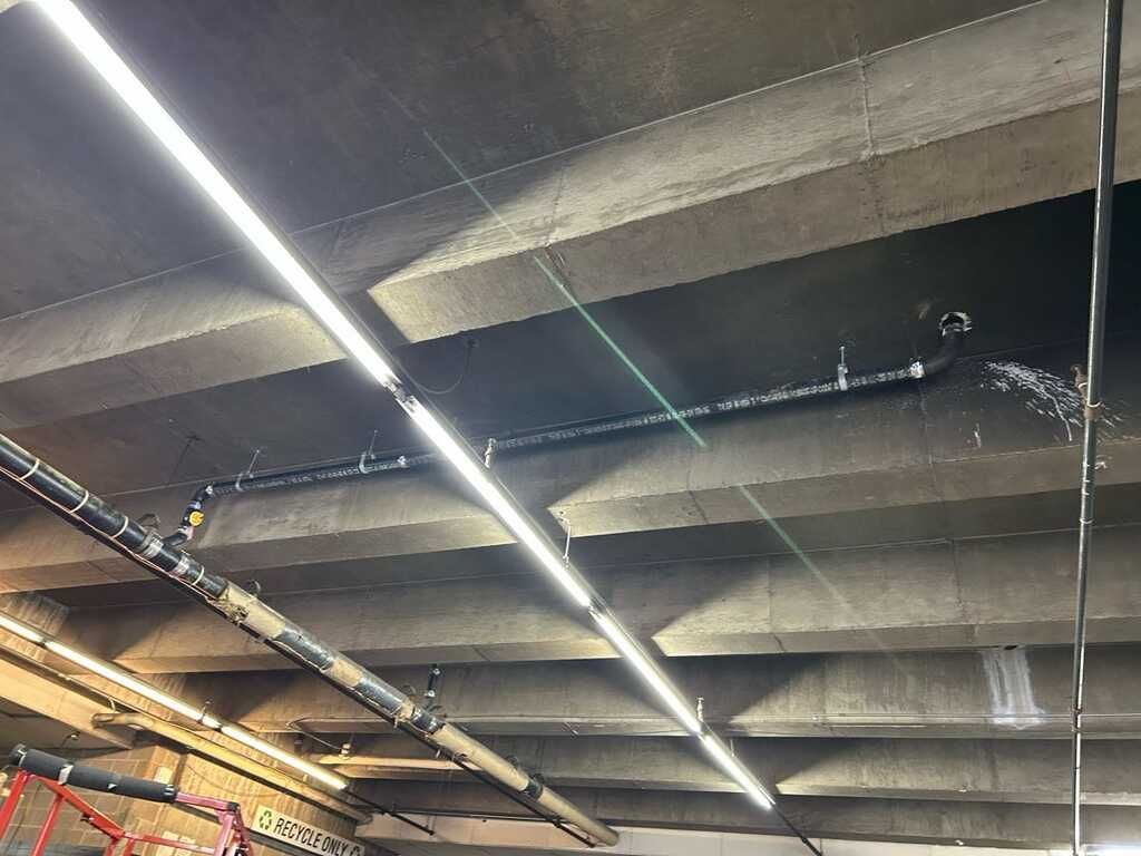 Ceiling with long fluorescent lights and exposed pipes in an industrial or parking garage setting
