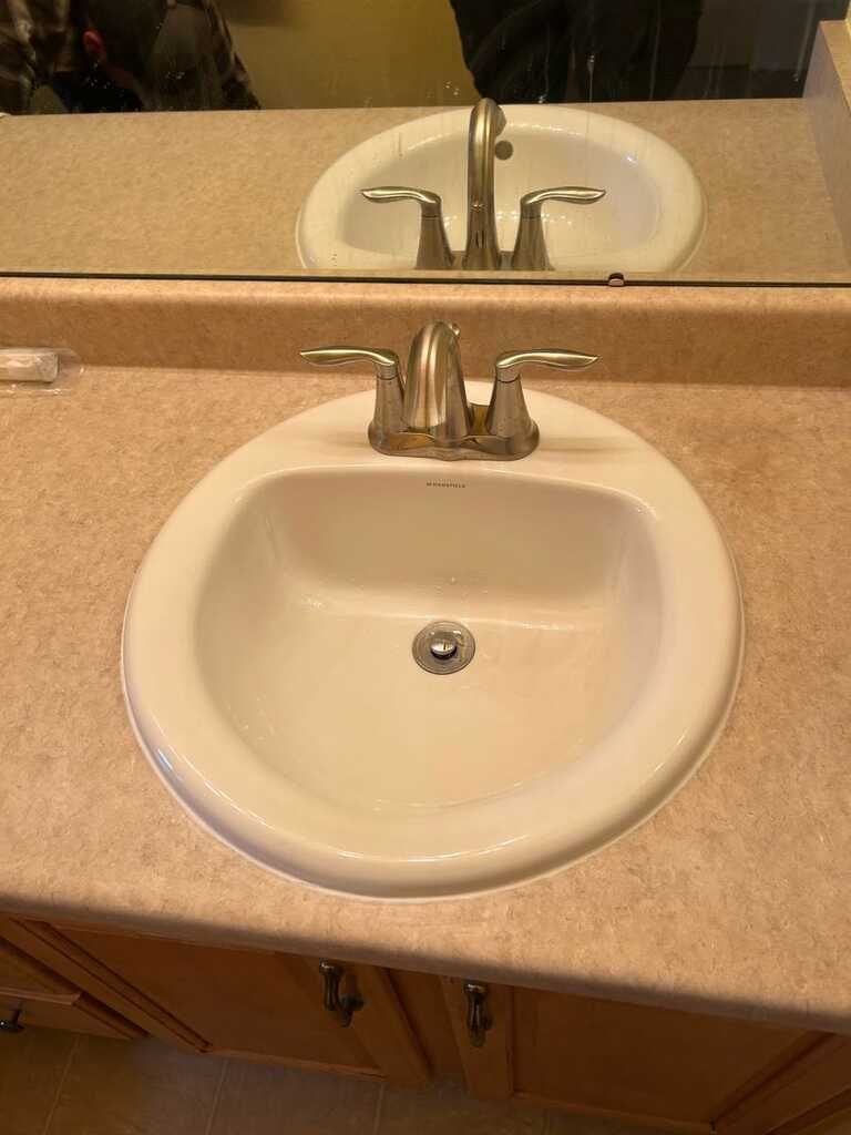 Small white bathroom sink with chrome faucet and beige countertop, seen with its reflection in the mirror.
