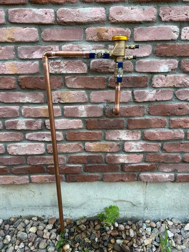 Copper pipe and brass valve mounted on a brick wall above gravel ground.