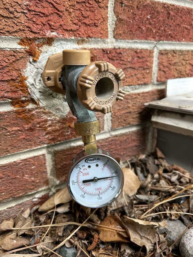 Outdoor water spigot with pressure gauge attached to a brick wall, surrounded by dry leaves.