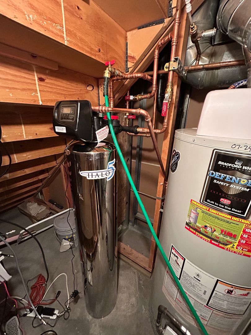Water softener and metal water tank in a utility closet with exposed pipes and a green hose.