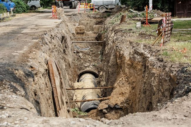 Roadside excavation with a large exposed pipe in a muddy trench and construction vehicles nearby