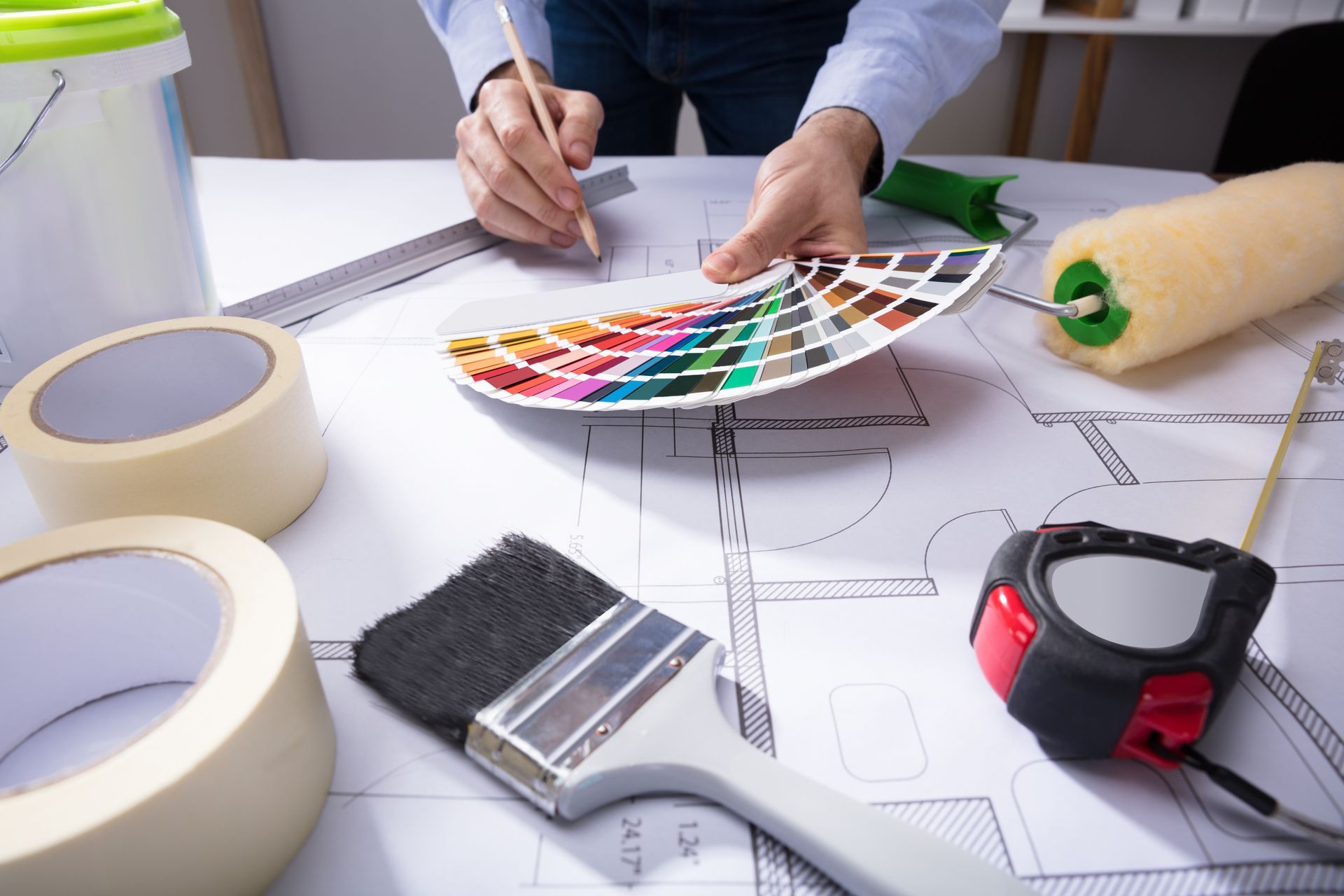 Hands choosing paint colors from a swatch fan over architectural drawings and tools on a desk