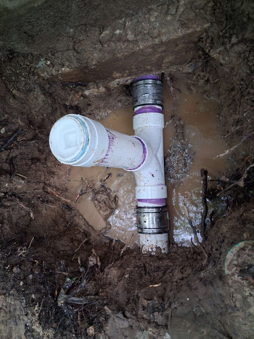 Black drainage pipe runs down a muddy hillside in a wooded area under an overcast sky