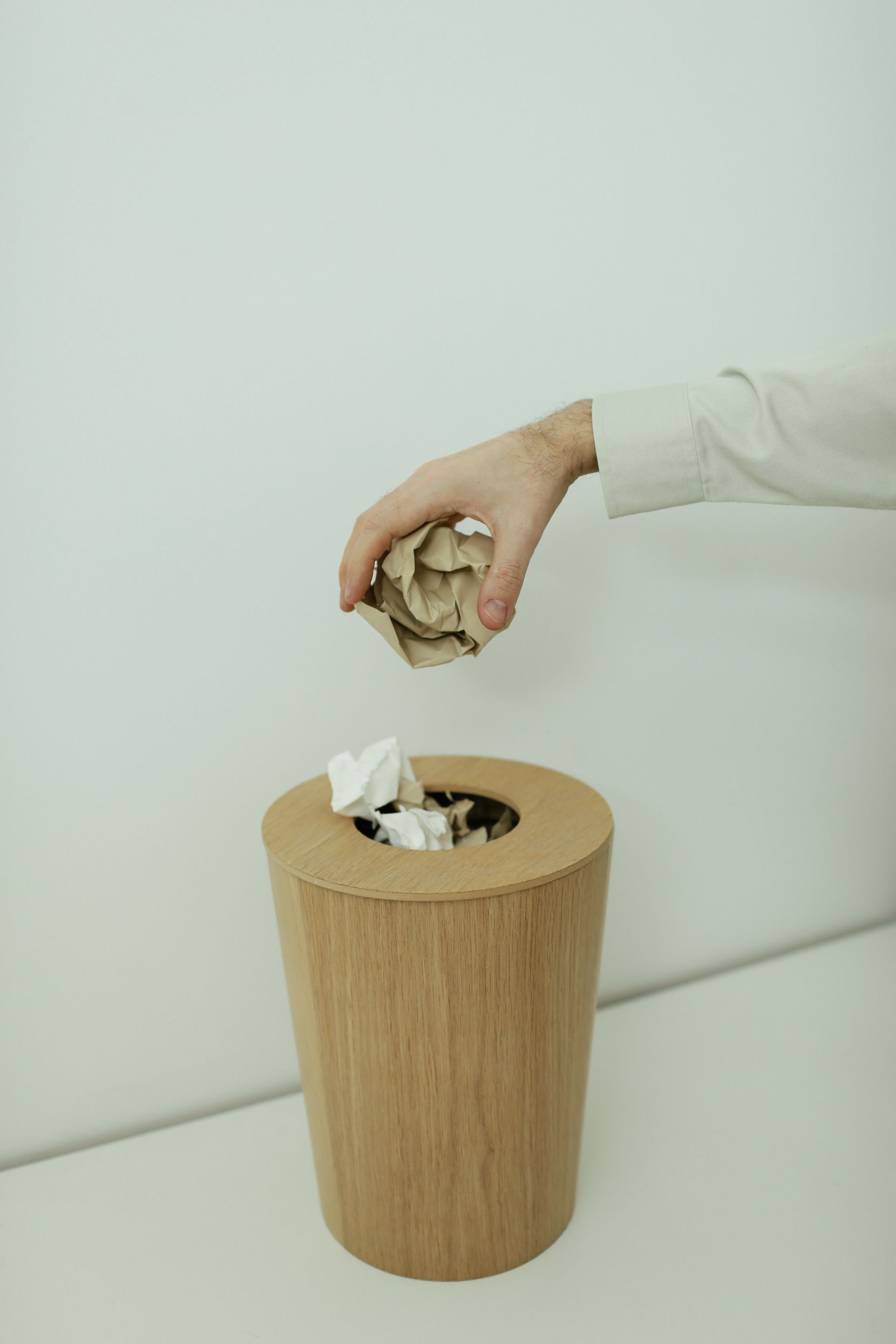 Hand dropping crumpled paper into a wooden trash bin against a white wall