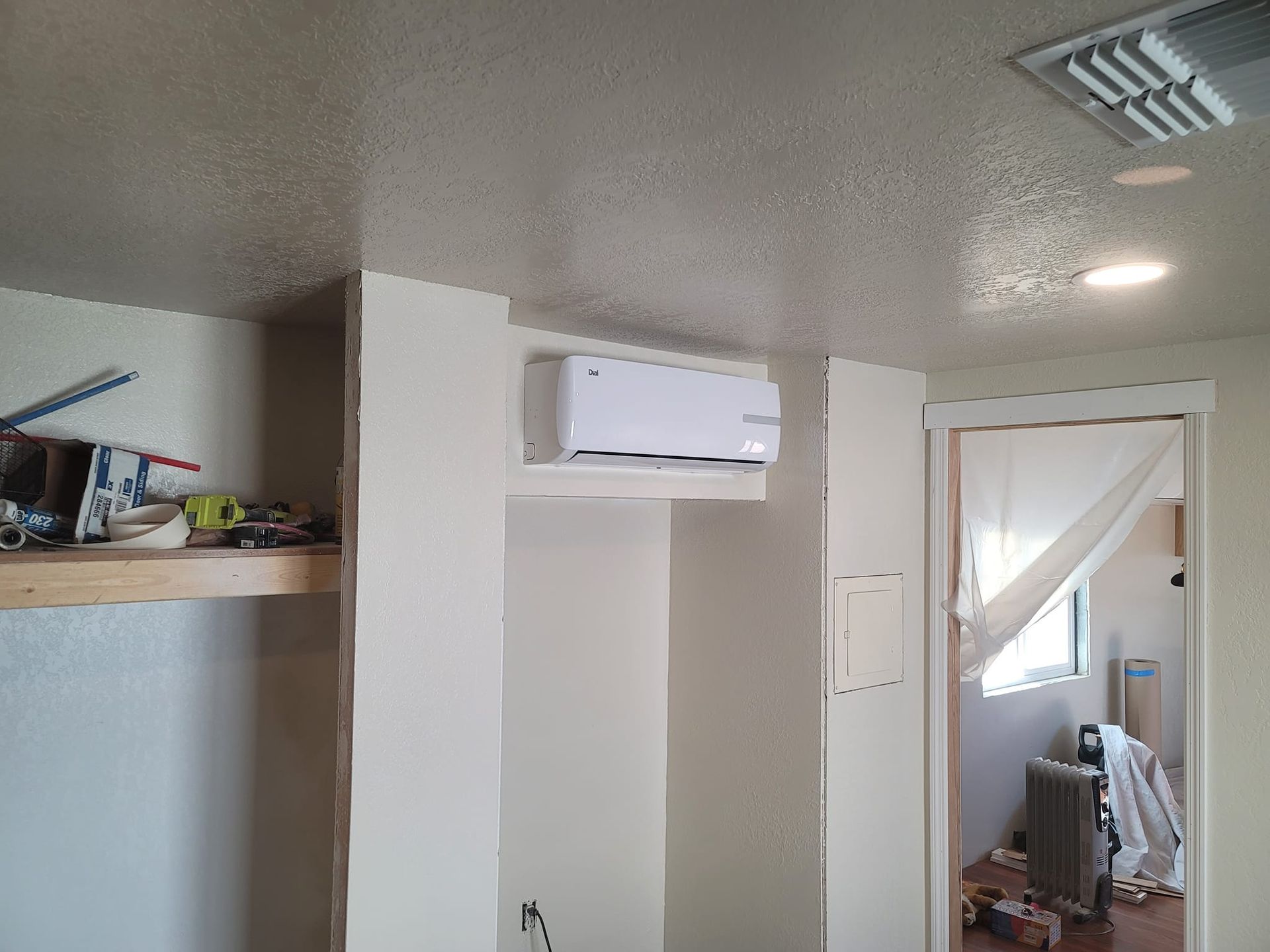 White wall-mounted air conditioner unit in a room with white walls, a partially visible doorway, and a shelf with items.