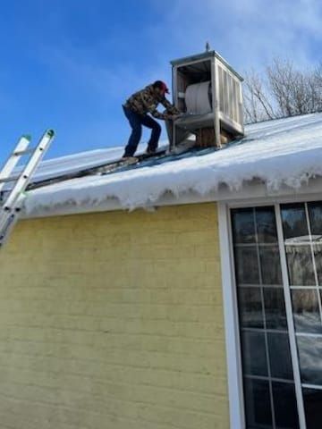 Person on snowy roof adjusting a rooftop vent under a clear blue sky. A ladder rests on the roof.