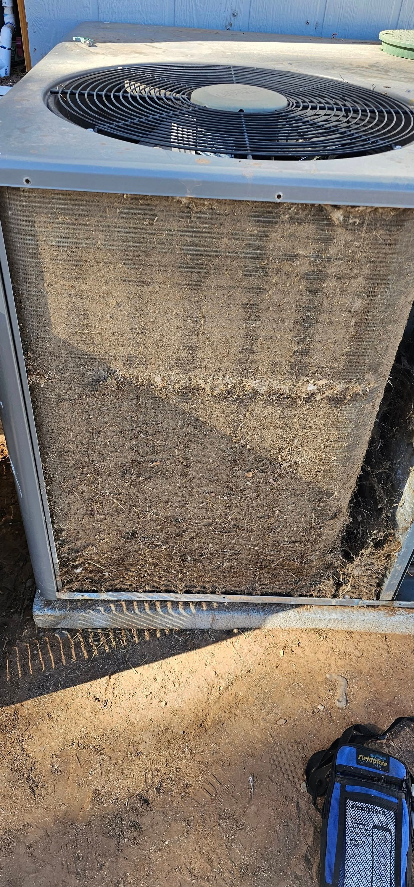An air conditioner unit's fins are covered in debris, sitting on a sandy ground with a tool bag nearby.