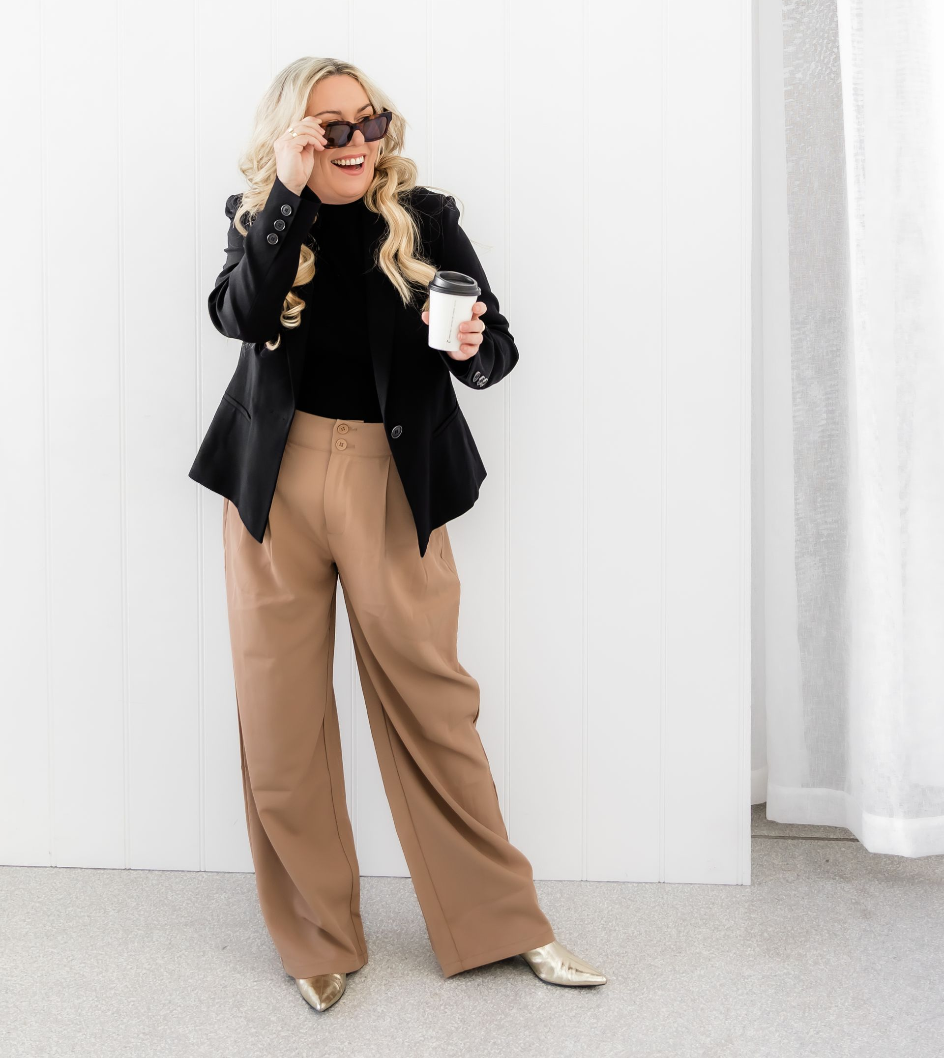 Woman wearing sunglasses, holding coffee, leans against white wall, wearing black top, blazer, tan pants, gold shoes.