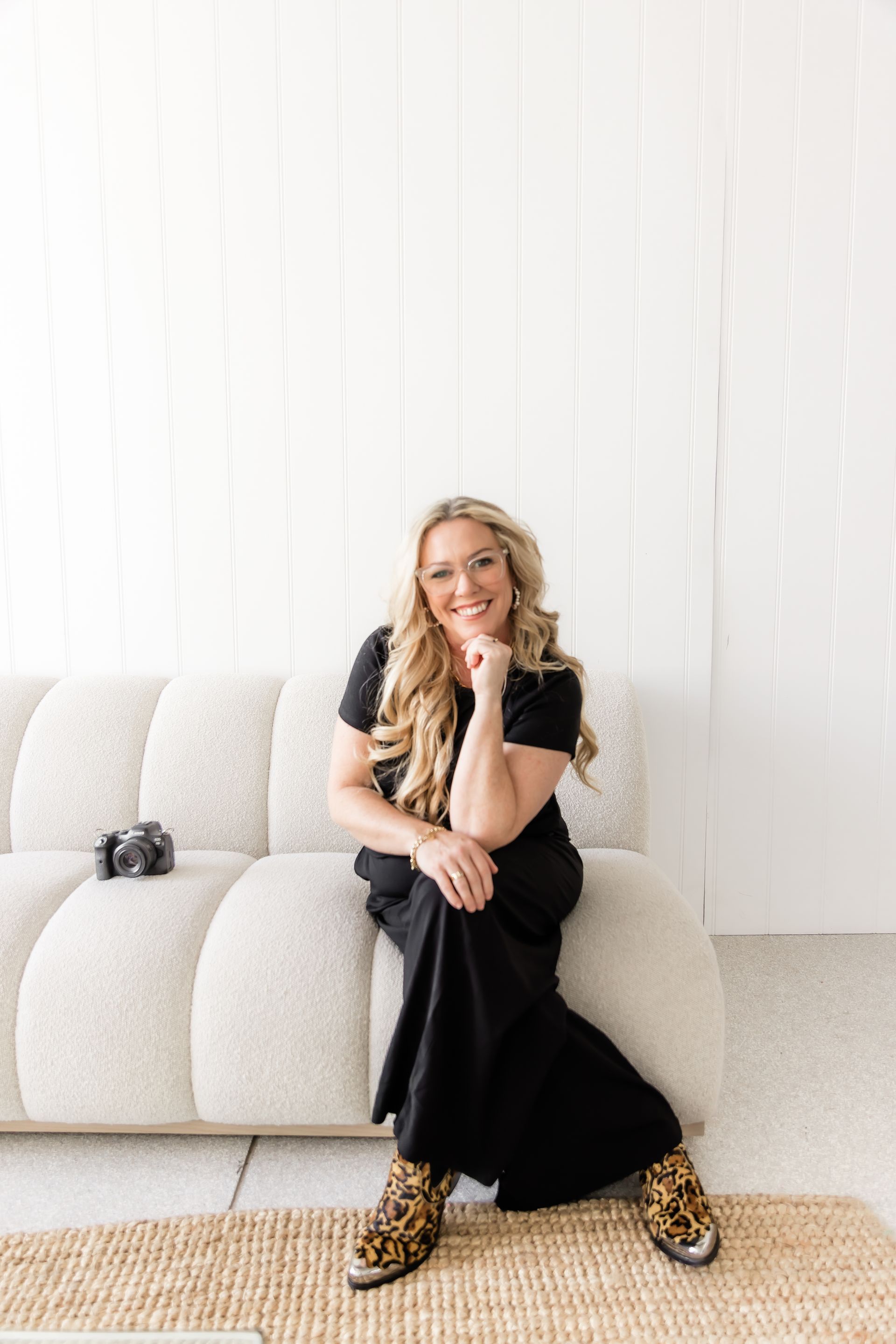 Woman in black dress and leopard print boots sits on a cream-colored couch, smiling with hand on chin.