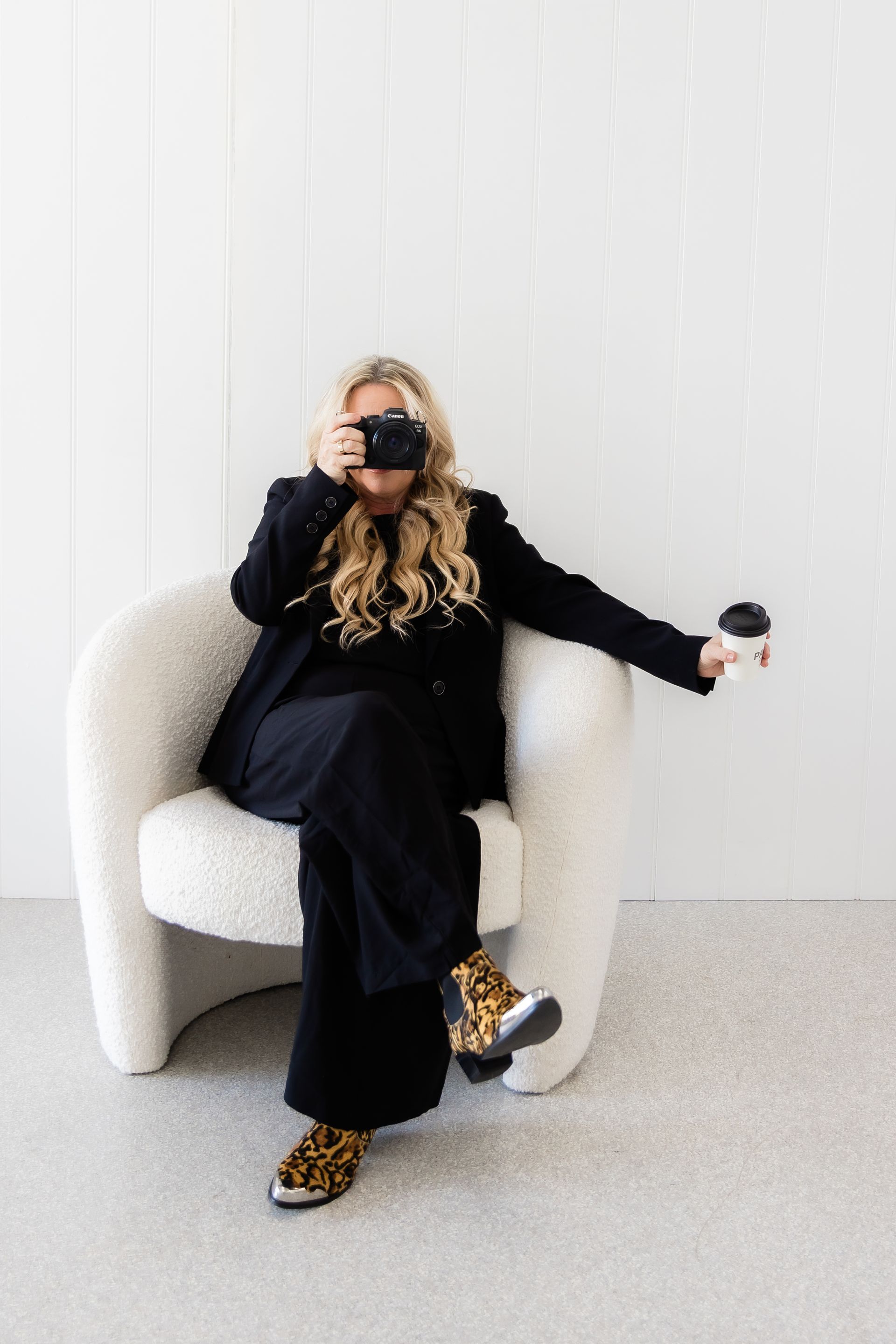 Woman in black suit, leopard print shoes, takes photo while seated, holding coffee. White setting.