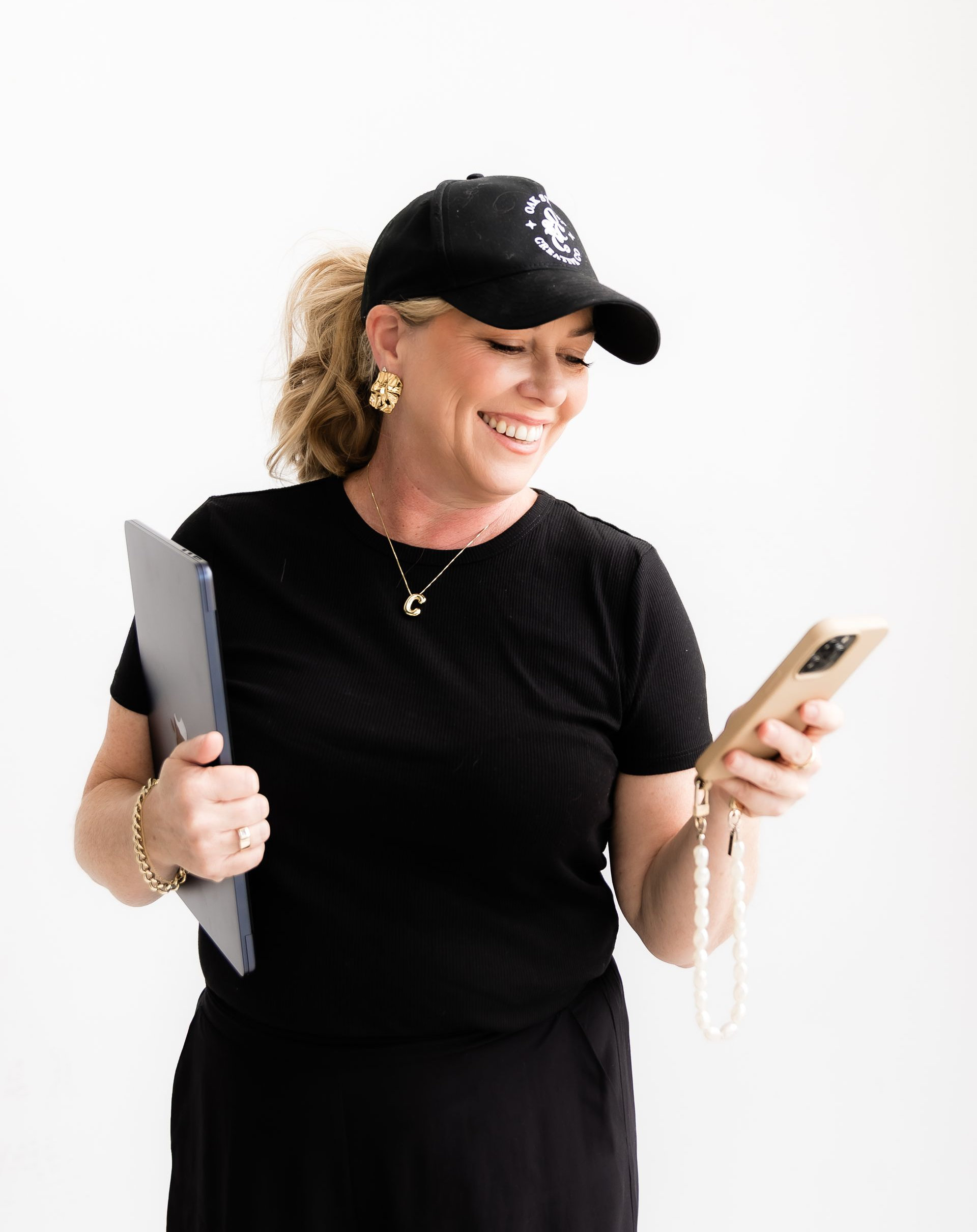 Woman in black holding laptop and phone, smiling. Wearing a cap, gold jewelry, against a white background.