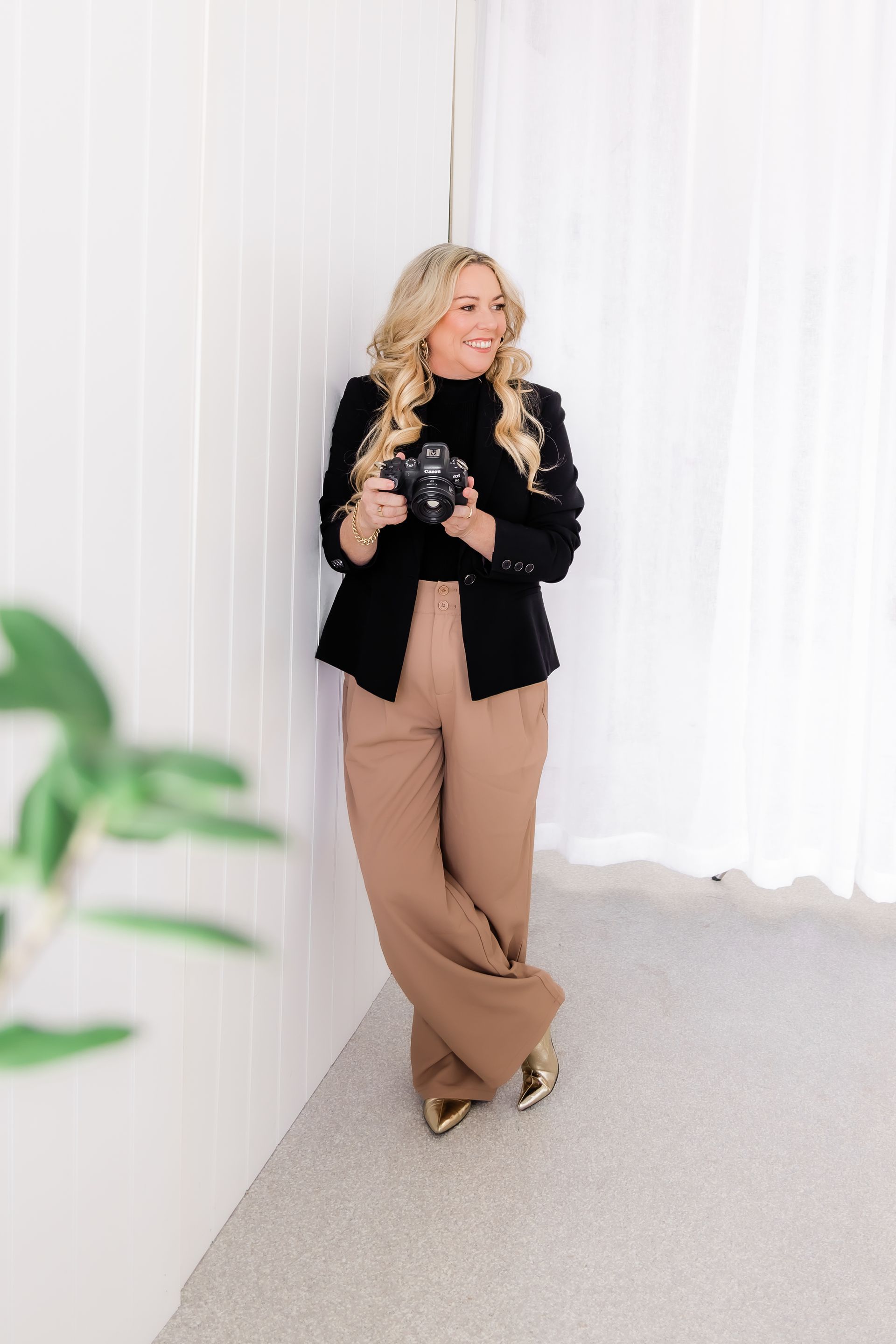 Blonde woman in black blazer and tan pants leans against a white wall, holding a camera, smiling.
