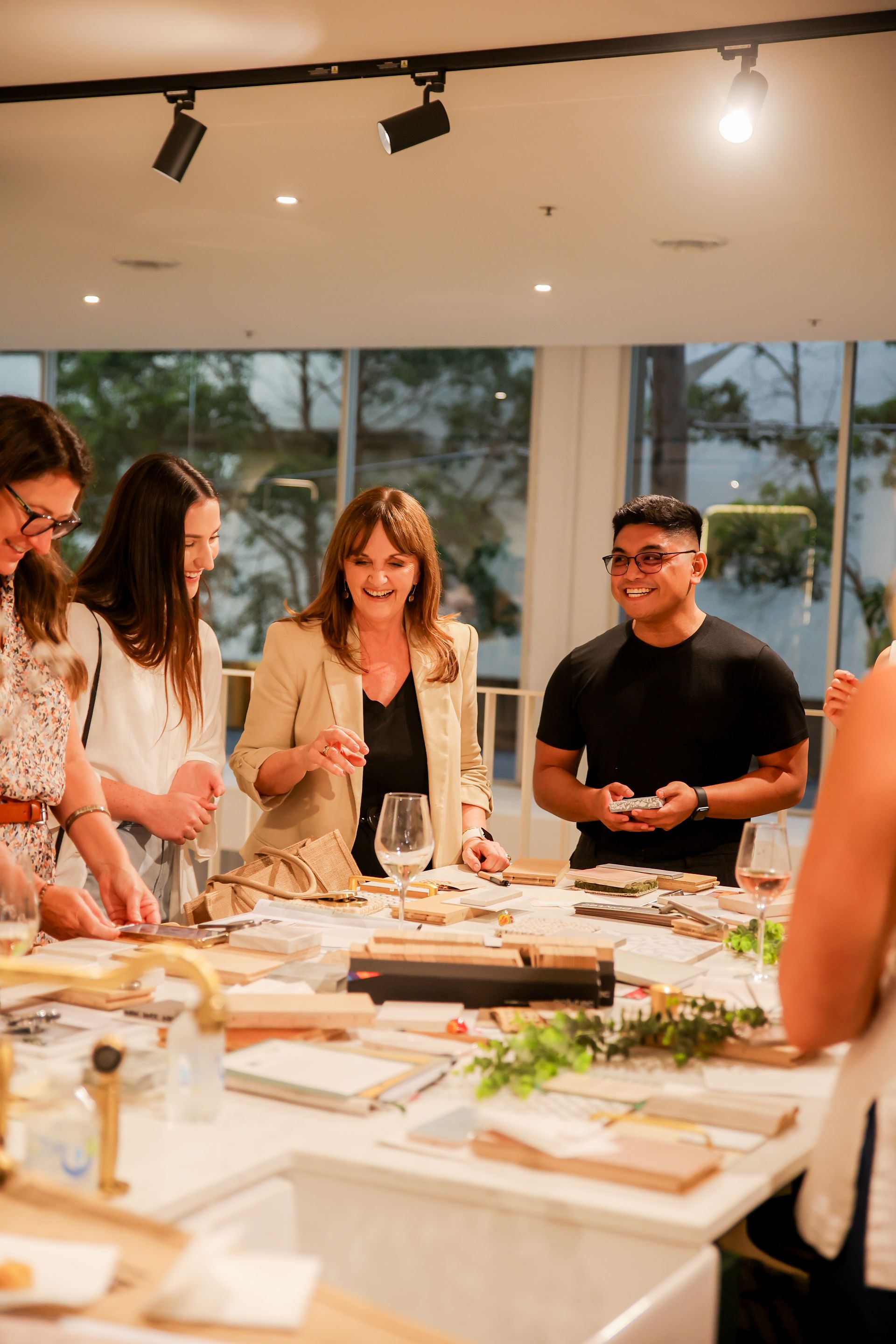 People at a workshop gathered around a table with items, smiling. Bright indoor setting.
