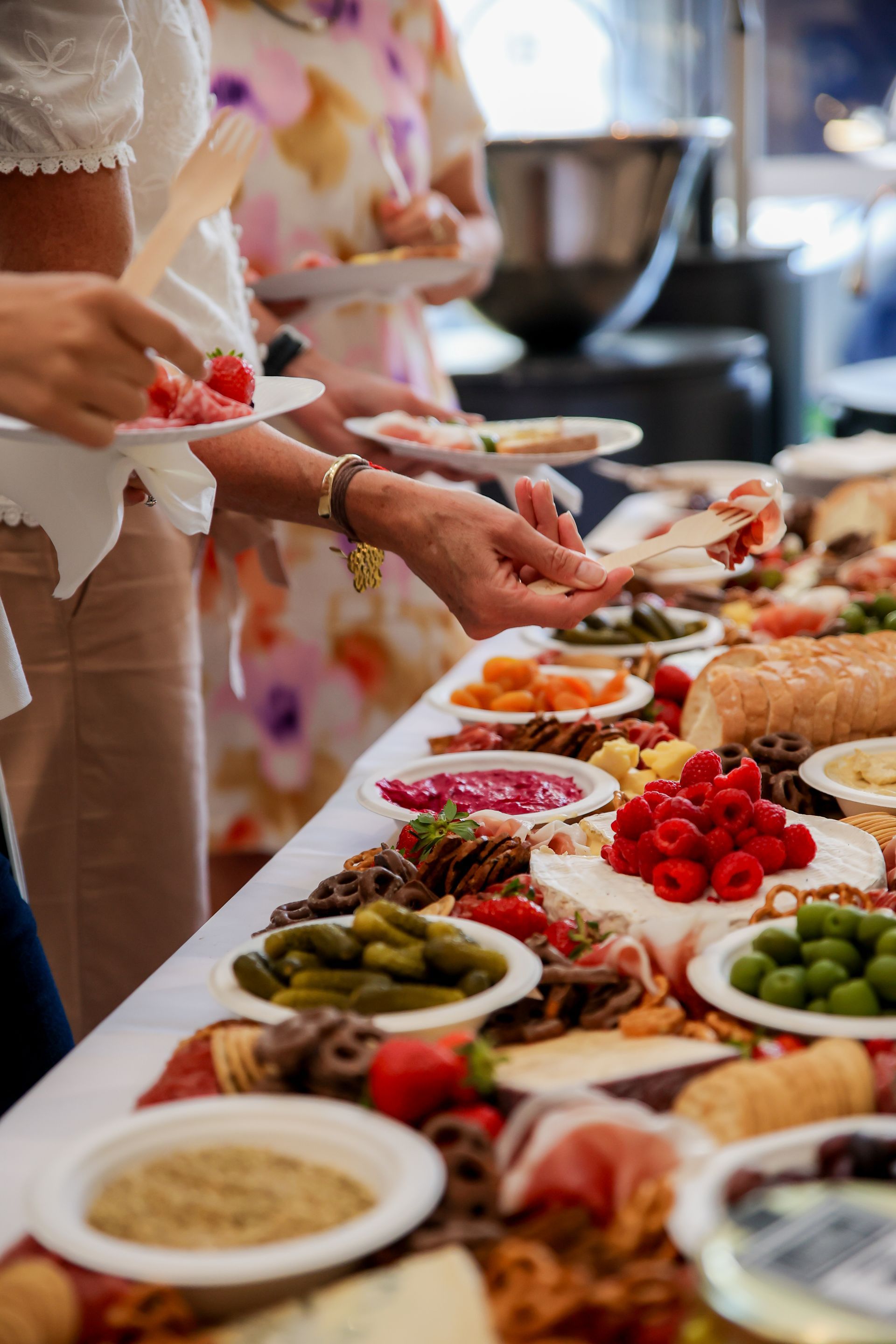 People serving themselves from a buffet table with various foods: fruits, dips, pickles.