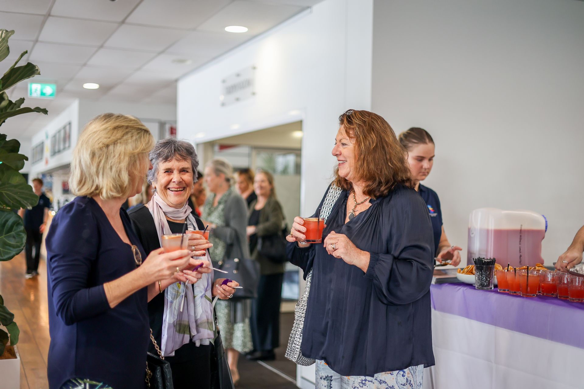 People socializing at an event, holding drinks, some smiling. Purple bar with refreshments in background.